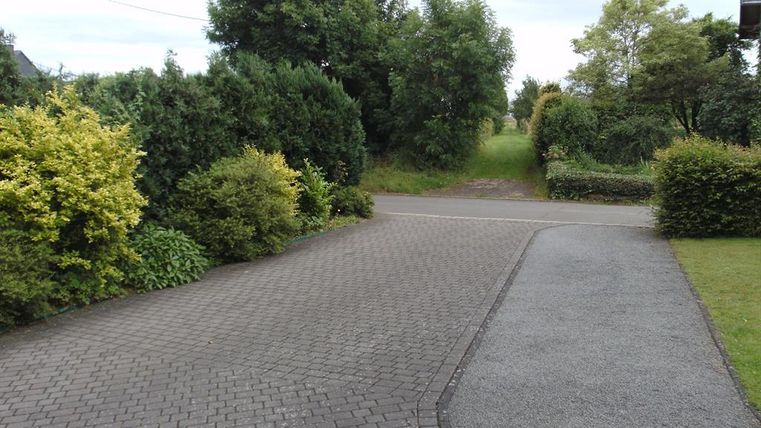 Ein gepflasterter Weg führt durch eine grüne Landschaft mit unterschiedlichen Sträuchern und Bäumen. Im Hintergrund ist ein schmaler Zugang zu einem Wiesenbereich sichtbar.