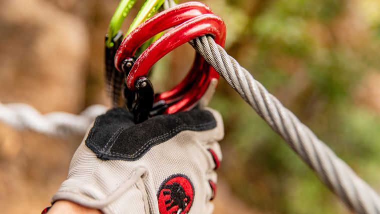 Ein Hand mit einem speziellen Handschuh hält mehrere Karabiner an einem Seil. Im Hintergrund sind unscharfe, grüne Bäume zu sehen.