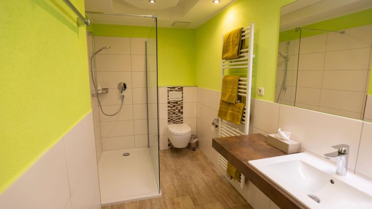 Ein modernes Badezimmer mit einer großen Dusche und einer hellgrünen Wandfarbe. Die Einrichtung umfasst ein Waschbecken, eine Toilette und Handtücher in Gelb.