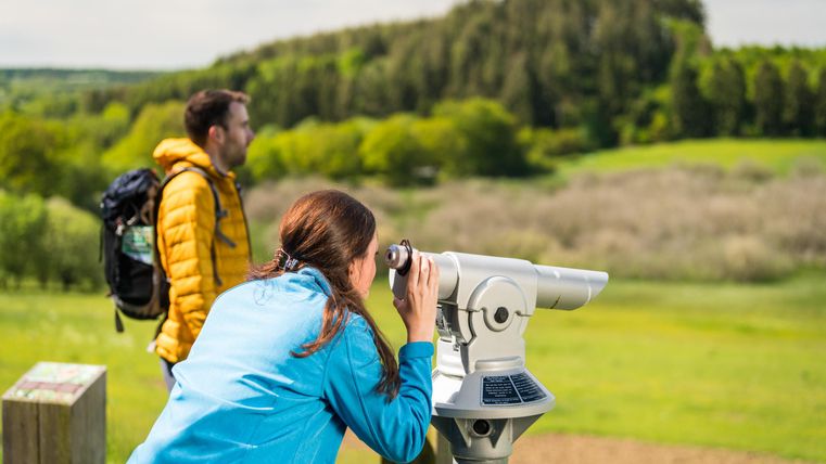Eine Frau schaut durch ein Fernglas und genießt die Aussicht auf eine grüne Landschaft. Im Hintergrund steht ein Mann mit einem Rucksack.