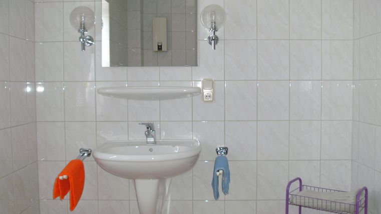 Ein einfaches Badezimmer mit weißen Fliesen und einem Waschbecken. Neben dem Waschbecken hängen zwei Handtücher und ein Spiegel ist an der Wand angebracht.
