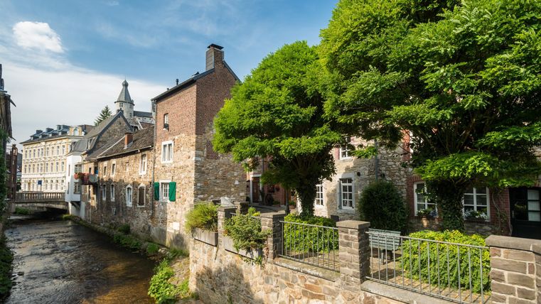 Eine malerische Szene mit historischen Häusern entlang eines ruhig fließenden Bachs. Dichte Bäume spenden Schatten und der Himmel ist klar und blau.