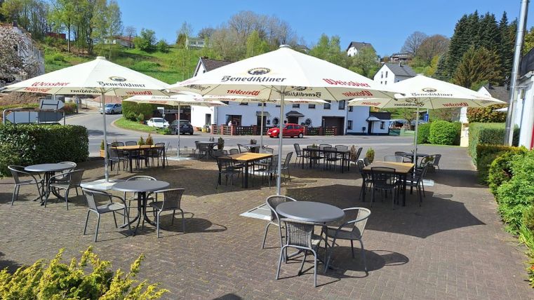 Een gezellige buitenruimte met tafel en stoelen onder grote parasols. Op de achtergrond zijn de groene heuvels en een restaurant zichtbaar.