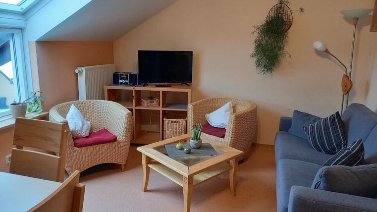 Een gezellige woonkamer met een helder dakraam. Er staan twee rieten stoelen, een grijze bank en een tafel in het midden.