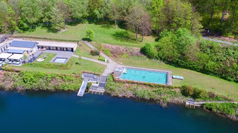 Eine ruhige Landschaft mit einem Schwimmbecken und einem klaren Gewässer. Umgeben von grünen Bäumen und Wiesen.