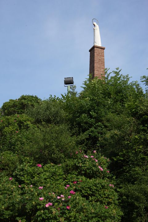 Mariensäule mit Marienstatue auf Backsteinsockel, umgeben von grünen Büschen und rosa Blumen, vor blauem Himmel.