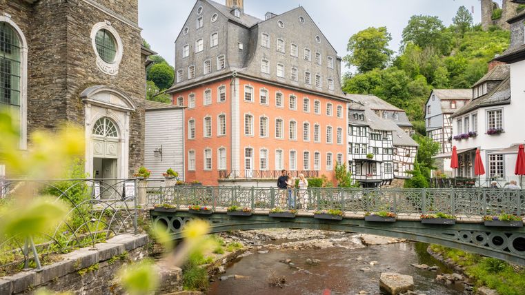 Eine malerische Stadtansicht mit bunten Häusern und einer kleinen Brücke über einen ruhigen Fluss. Im Hintergrund sind grüne Hügel und ein historisches Gebäude zu sehen.