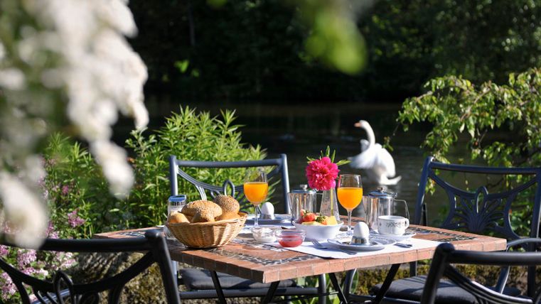An inviting outdoor table set with breakfast and fresh rolls. In the background, flowers and a calm water surface can be seen.