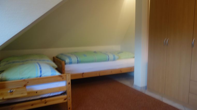 Een eenvoudige slaapkamer met twee eenpersoonsbedden en een houten kast. De muren zijn licht en de vloer is voorzien van tapijt.