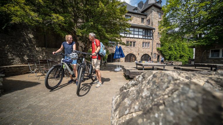 Zwei Radfahrer stehen auf einem Platz vor einem historischen Gebäude. Die Umgebung ist grün und einladend, ideal für Outdoor-Aktivitäten.