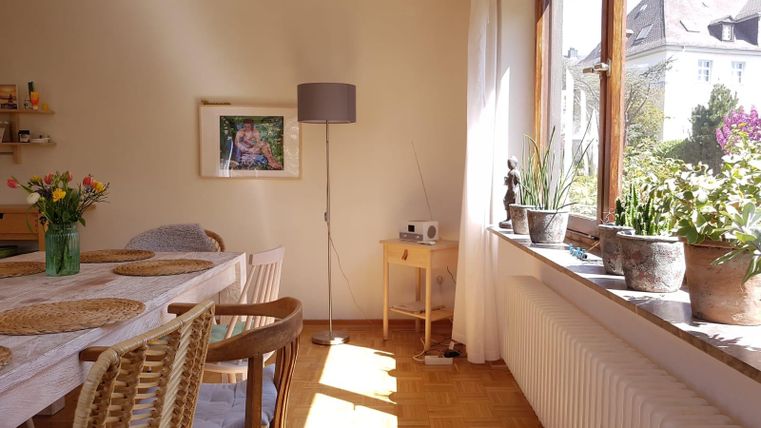 Een lichte woonkamer met een tafel, stoelen en een vloerlamp. Planten en bloemen staan op de vensterbank en er hangt een schilderij aan de muur.