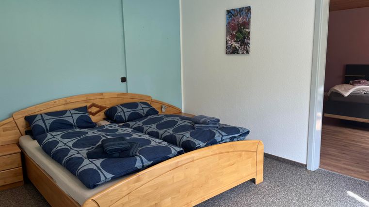 A modern bedroom with a wooden double bed and blue bedding. The walls are painted in soft colors, and there is a window that lets in natural light.