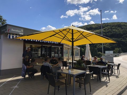 A cozy outdoor café with a large yellow sun umbrella. Guests are sitting at tables and enjoying the sun.
