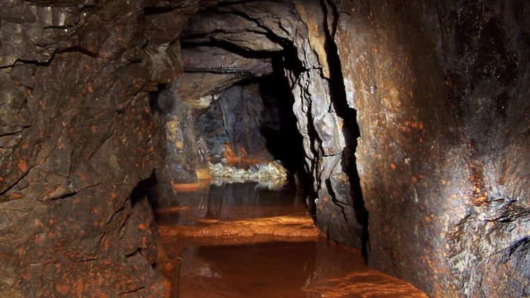 A dark tunnel with rocky walls and still, brownish water. In the background, faint light can be seen that highlights the structure of the rock.
