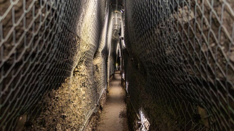 Een smalle gang met een bodem van aarde en wanden die door draadhekken zijn beveiligd. De verlichting zorgt voor een gedempte sfeer.
