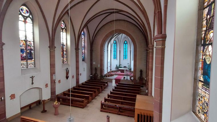 Een stille kerk met hoge plafonds en kleurrijke glas-in-loodramen. De banken staan netjes op een rij en het altaar is goed zichtbaar.
