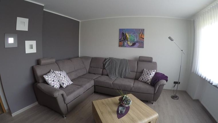 A modern living room with a beige L-shaped couch and decorative cushions. The room features a wooden table and an elegant floor lamp, complemented by a colorful picture on the wall.