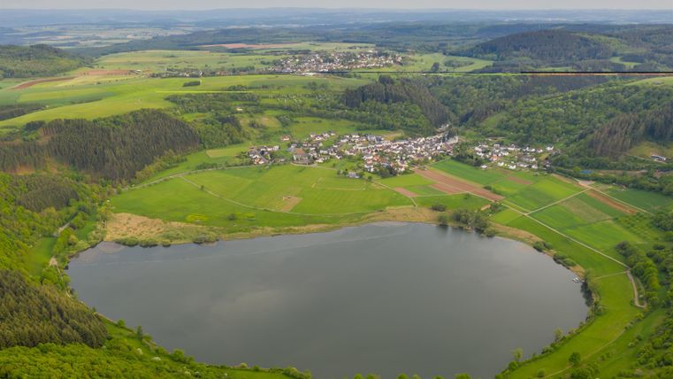 Een schilderachtig landschap met een rustige meer en weelderig groen. Op de achtergrond rijzen zachte heuvels op en een klein dorp is zichtbaar.
