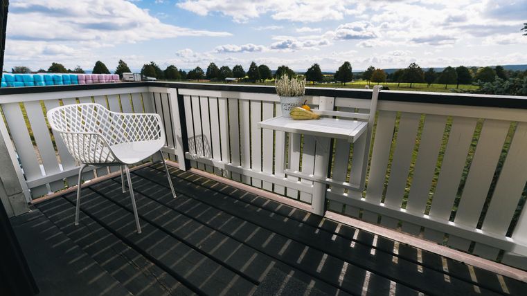Een gezellige balkon met een witte stoel en een klein tafeltje. Op de achtergrond strekt zich een groene landschap uit onder een gedeeltelijk bewolkte lucht.