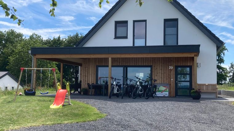 Ein modernes Haus mit einer Holzveranda und einem Spielplatz im Vordergrund. Fahrräder stehen vor dem Eingang bereit, um genutzt zu werden.