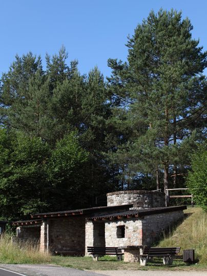 An old lime kiln made of stone stands in a forest environment. Benches are located in front of the kiln. The sky is clear and blue.