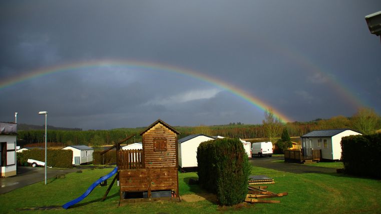 Ein Spielplatz mit einem Holzhäuschen und einer Rutsche. Im Hintergrund leuchtet ein Regenbogen über einem Campingplatz.