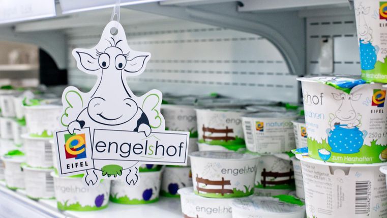 Ein Regal mit verschiedenen Joghurtbechern und einem Schild mit einer lustigen Kuh. Das Produkt stammt von der Marke "engels hof".