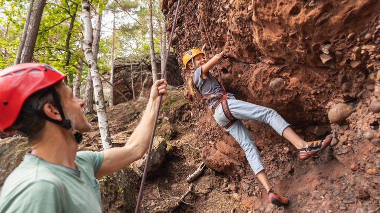 Een klimmer hangt aan een rotswand terwijl een persoon hem beveiligt. Beiden dragen helmen en zijn omgeven door bomen.