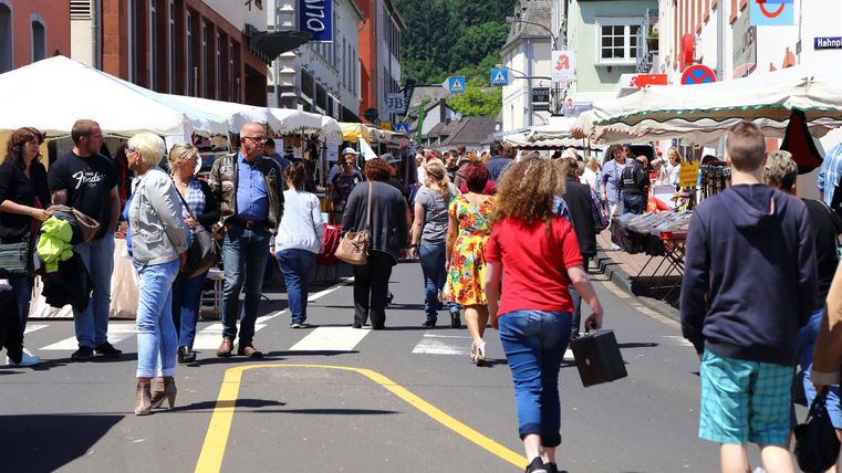 Een drukke marktdag op een straat, omringd door kraampjes en veel mensen. De sfeer is levendig en vriendelijk, met kleurrijke kleding en ingekochte goederen.