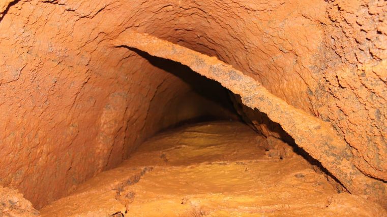 A narrow tunnel with reddish-brown walls and a rough surface. The floor is made of a mineral material that appears to be wedged between the walls.