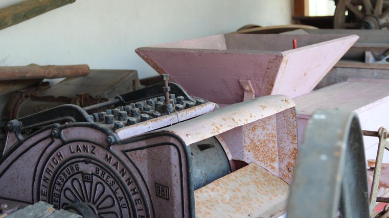 Eine alte Landmaschine aus Metall, rostig und abgenutzt. Im Hintergrund sind weitere landwirtschaftliche Werkzeuge zu sehen.