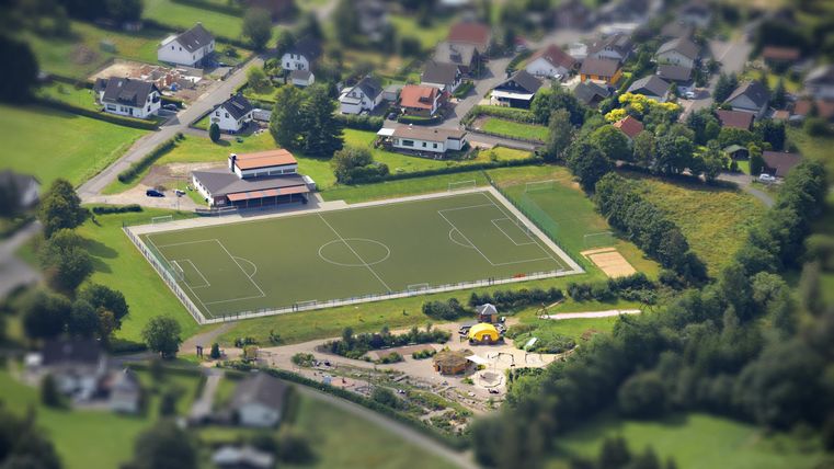 Ein Fußballplatz umgeben von Wohngebieten und grünen Wiesen. Im Vordergrund ist ein Spielplatz zu sehen.
