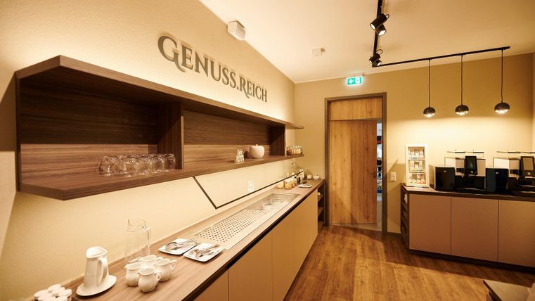 Ein moderner Raum mit einem langen Tisch, ausgestattet mit Gläsern und Kaffeemaschinen. Die Wände sind in warmen Tönen gehalten, und es gibt eine einladende Atmosphäre.