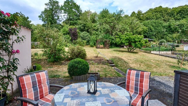 Eine gemütliche Terrasse mit einem runden Tisch und zwei Stühlen. Im Hintergrund sieht man einen schönen Garten mit Bäumen und Sträuchern.
