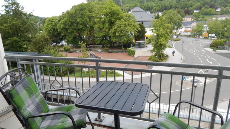A balcony with two chairs and a small table. In the background, trees and a street view can be seen.