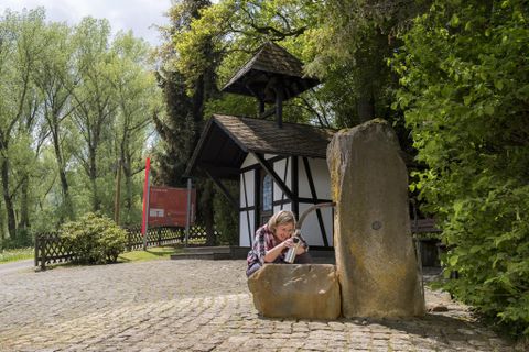 Ein Junge sitzt an einem Wasserbrunnen und schaut auf sein Handy. Im Hintergrund ist ein kleines, traditionelles Fachwerkgebäude und viel grüne Natur.