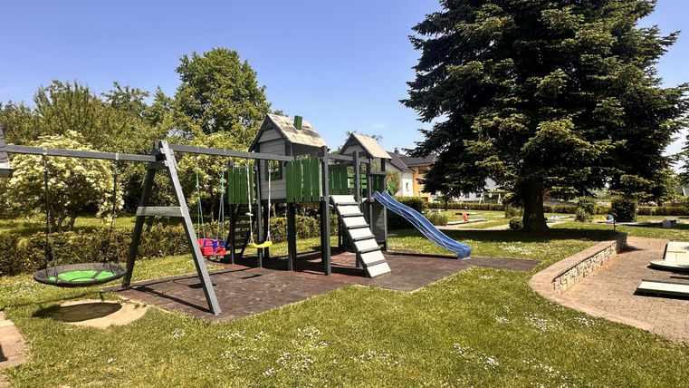Ein Spielplatz mit einer Rutsche, einer Schaukel und einem Klettergerüst. Umgeben von grünem Gras und Bäumen bei strahlend blauem Himmel.