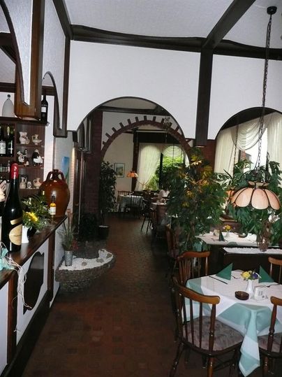 Cozy interior of a pizzeria with wooden furniture, plants, and set tables. An arch leads to additional seating areas.