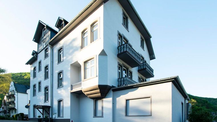 Ein modernes, weißes Gebäude mit Balkonen und großen Fenstern. Es befindet sich in einer ruhigen Umgebung mit grünen Hügeln im Hintergrund.