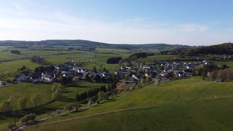 A picturesque landscape with gentle hills and a small village. The village is surrounded by green fields and lies under a clear blue sky.