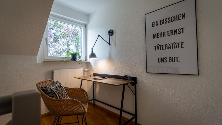 Ein gemütlicher Arbeitsbereich mit einem Schreibtisch und einem bequemen Stuhl. Das Fenster ist geöffnet und lässt Tageslicht herein.