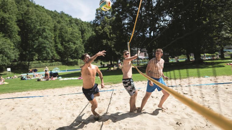 Drei Männer spielen Beachvolleyball an einem sonnigen Tag. Die Umgebung ist grün und einladend, mit vielen Menschen im Hintergrund.