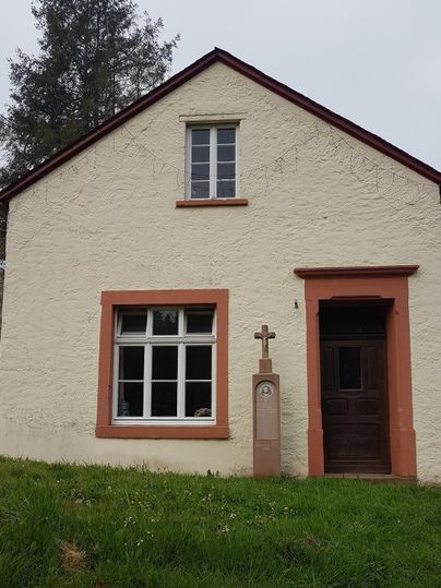 Ein kleines, charmantes Haus mit einem hellen Anstrich und einem roten Akzent. Vor der Tür steht ein Kreuz und die Umgebung ist grün und ländlich.