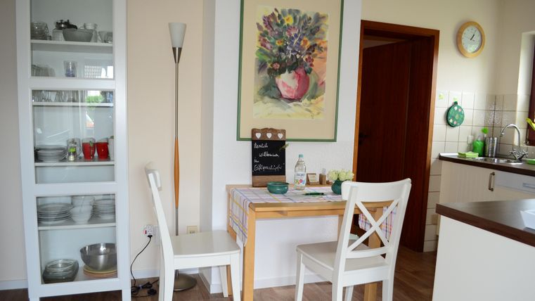 A cozy dining area with a small table and two chairs. A colorful flower picture hangs on the wall, and in the corner stands a glass shelf.