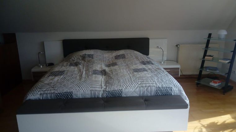 A modern bedroom with a large bed and a cozy blanket. Next to it is a glass bedside table and a subtly lit lamp.