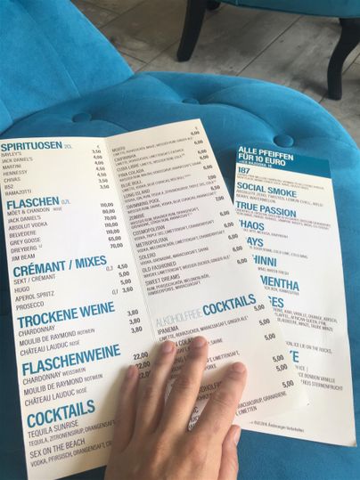 Ein Getränkemenü mit verschiedenen Optionen wie Weinen und Cocktails. Eine Hand hält das Menü auf einem blauen Sitz.