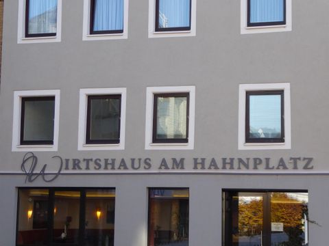 Ein modernes Wirtshaus mit der Aufschrift "Wirtshaus am Hahnplatz". Die Fassade zeigt mehrere Fenster in einer hellgrauen Wand.