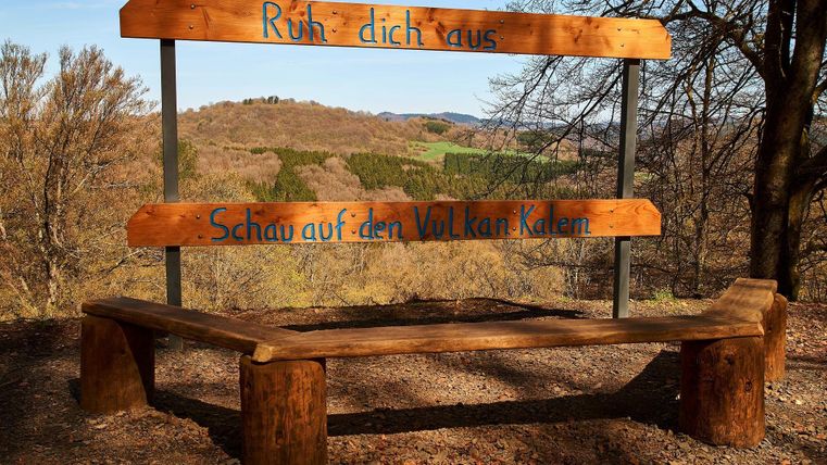 Eine Holzbank mit der Aufschrift „Ruh dich aus“ und „Schau auf den Vulkan Kalem“. Im Hintergrund sind sanfte Hügel und Baumlandschaft zu sehen.