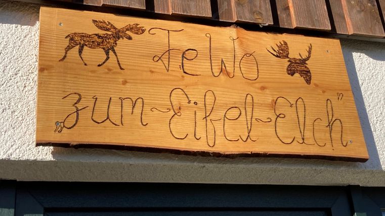 Ein Holzschild mit der Aufschrift „Ferienwohnung zum Eifel-Elch“ und zwei eingravierten Elchen. Das Schild hängt über einer Tür und vermittelt eine gemütliche Atmosphäre.