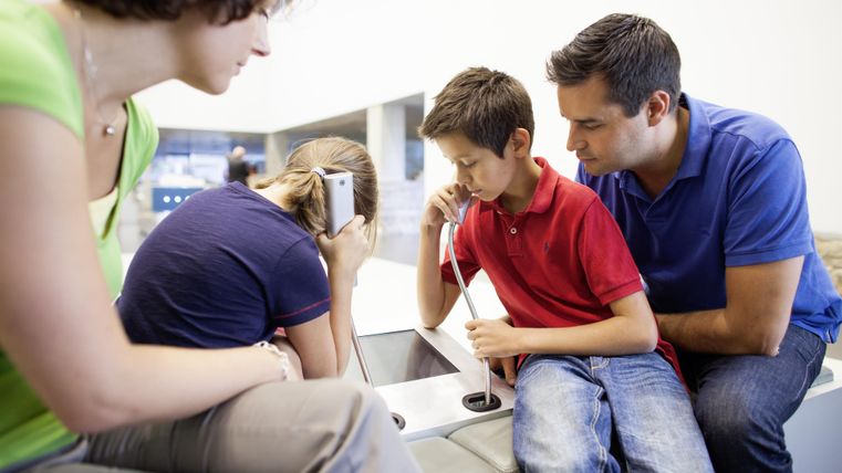 Eine Familie sitzt an einem Tisch und interagiert mit technischen Geräten. Die Kinder hören etwas über Kopfhörer, während die Erwachsenen sie beobachten.